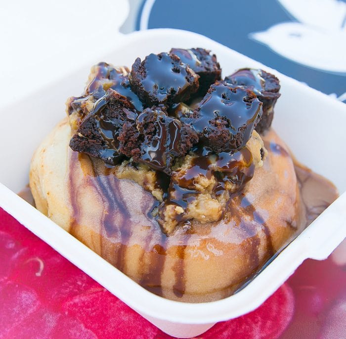photo of a cinnamon roll topped with Chocolate frosting, cookie dough, brownies, and chocolate syrup drizzle