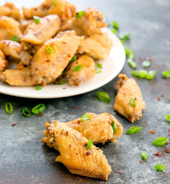 close up of two baked salt and pepper wings with a plate of them in the background