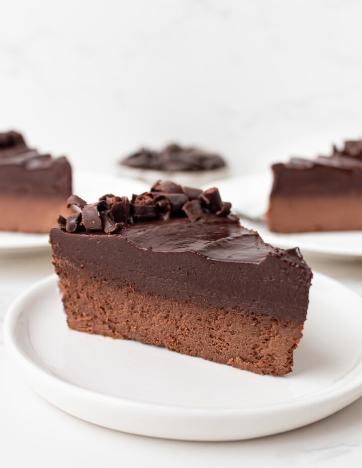 A slice of chocolate cake with a chocolate ganache frosting and chocolate curls on a small white plate