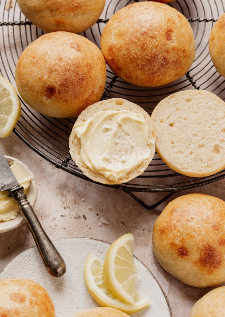 Golden-brown bread rolls cooling on a wire rack, with one roll sliced open and spread with butter.