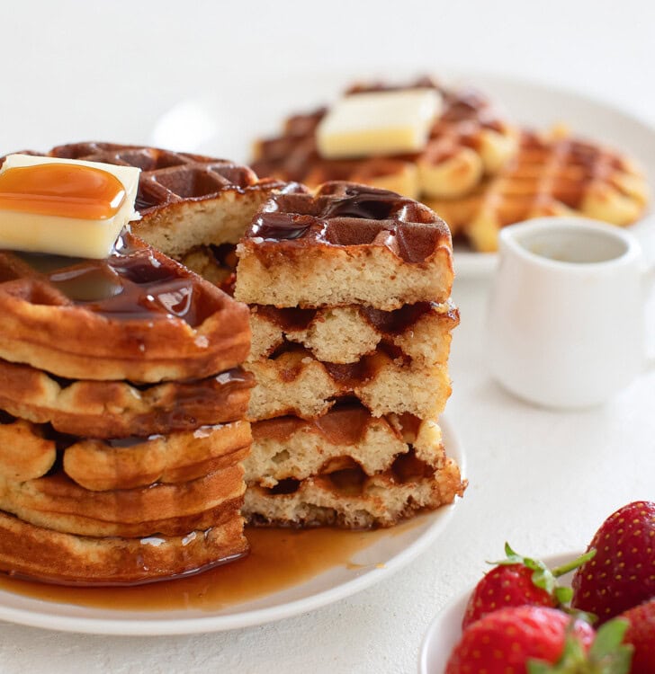 Stack of golden brown keto waffles topped with butter and syrup next to sliced waffles showing a fluffy interior